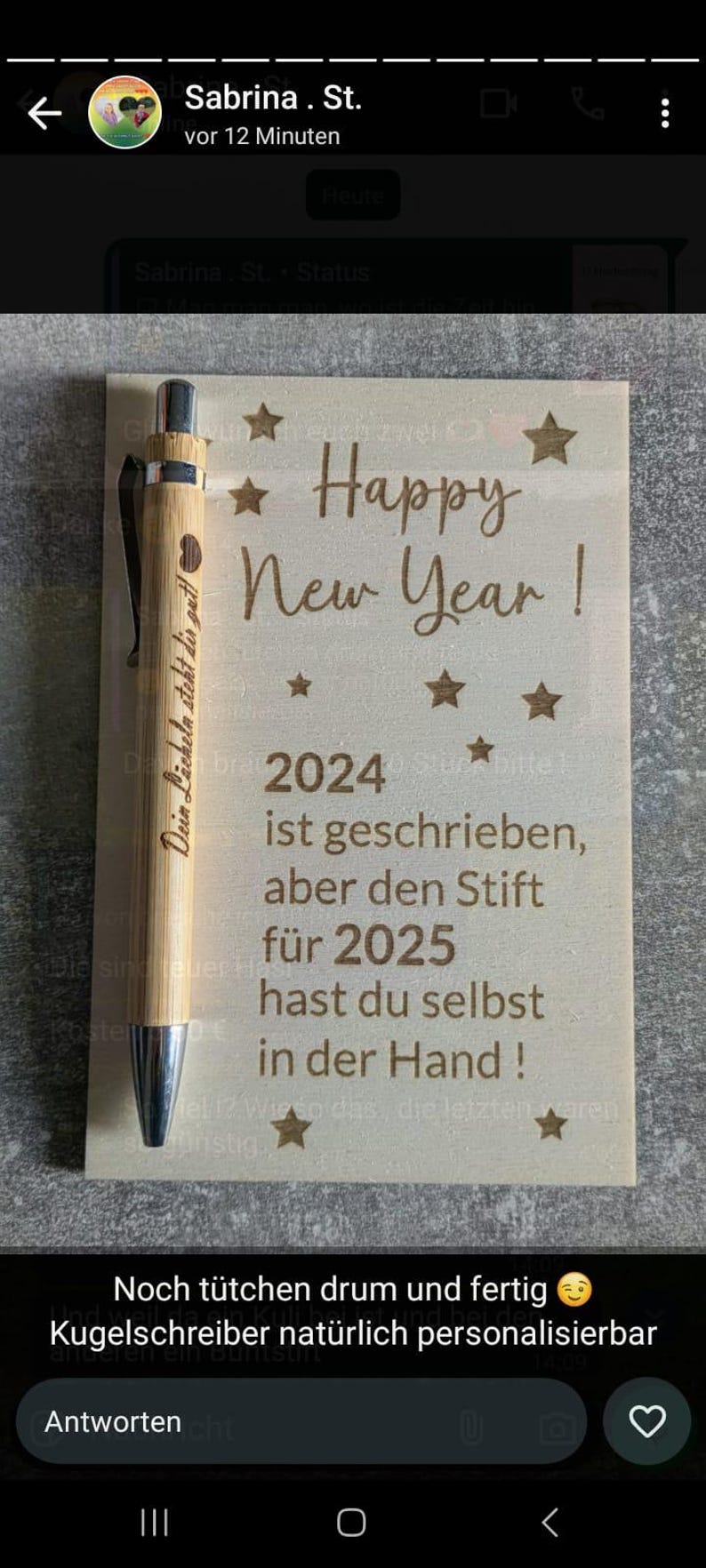 May include: A wooden sign with the words "Happy New Year!" and "2024" engraved, accompanied by star designs. A bamboo pen with a silver tip and the words "Dein L&auml;cheln steht dir gut!" is placed on the sign.