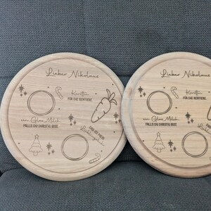 May include: Two round wooden serving trays with engraved designs for Santa Claus. Each tray has a space for milk, cookies, carrots, and a note for Santa. The text on the trays reads "Lieber Nikolaus" which is German for "Dear Santa".