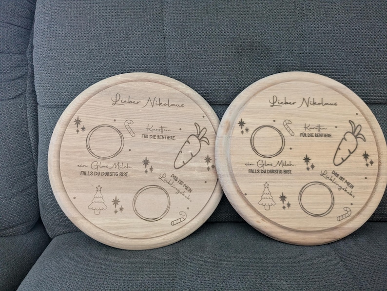 May include: Two round wooden serving trays with engraved designs for Santa Claus. Each tray has a space for milk, cookies, carrots, and a note for Santa. The text on the trays reads "Lieber Nikolaus" which is German for "Dear Santa".