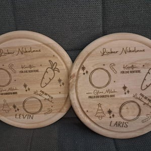 May include: Two round wooden plates with engraved designs for Santa Claus. The plates have a carrot, candy cane, a glass of milk, and a Christmas tree. One plate says "Lieber Nikolaus" and "Levin" and the other says "Lieber Nikolaus" and "Laris".