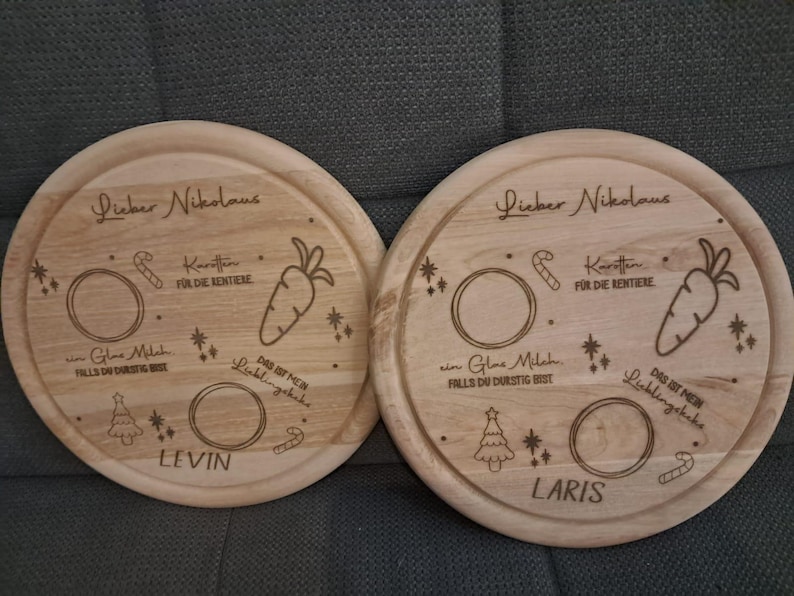 May include: Two round wooden plates with engraved designs for Santa Claus. The plates have a carrot, candy cane, a glass of milk, and a Christmas tree. One plate says "Lieber Nikolaus" and "Levin" and the other says "Lieber Nikolaus" and "Laris".