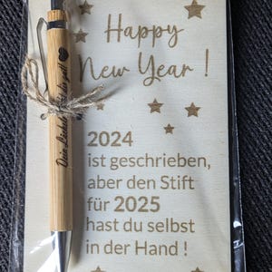 May include: A bamboo pen tied with twine, presented with a wooden card. The card reads "Happy New Year!" and includes the year 2024, with a message in German. The pen has a silver tip and a heart symbol.