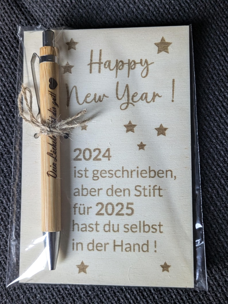 May include: A bamboo pen tied with twine, presented with a wooden card. The card reads "Happy New Year!" and includes the year 2024, with a message in German. The pen has a silver tip and a heart symbol.
