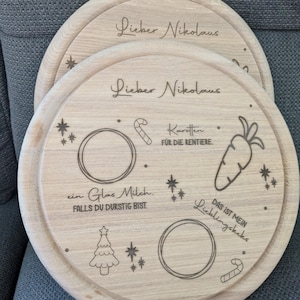 May include: A wooden round serving tray with a laser engraved design for Santa Claus. The tray features a Christmas tree, stars, candy canes, a glass of milk, and a carrot. The text on the tray reads "Lieber Nikolaus" and "Das ist mein Lieblingskeks".