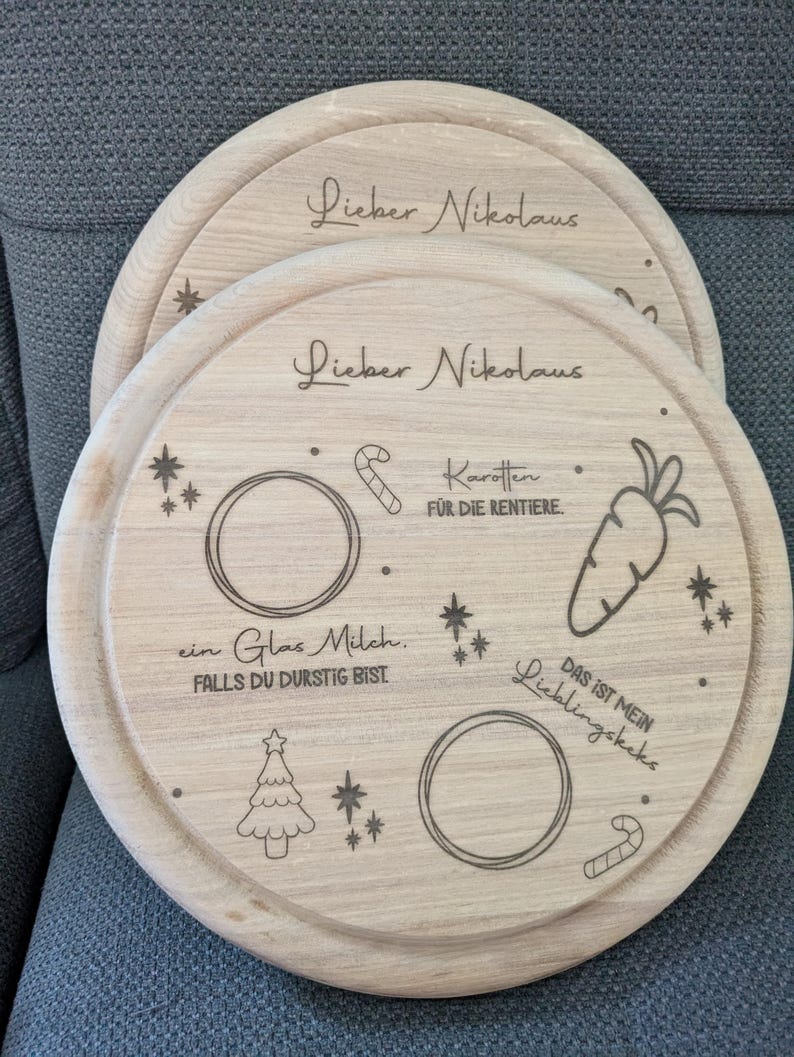 May include: A wooden round serving tray with a laser engraved design for Santa Claus. The tray features a Christmas tree, stars, candy canes, a glass of milk, and a carrot. The text on the tray reads "Lieber Nikolaus" and "Das ist mein Lieblingskeks".