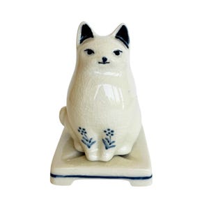 May include: A ceramic cat figurine in a seated position, with a cream-colored body and black accents on the ears and eyes. The figurine has a crackle glaze finish and is decorated with blue floral motifs on the base.