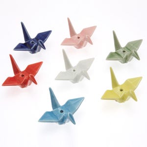 May include: Set of seven ceramic origami cranes in various colors, including blue, red, pink, white, green, and yellow. Each crane has a small hole in the center.