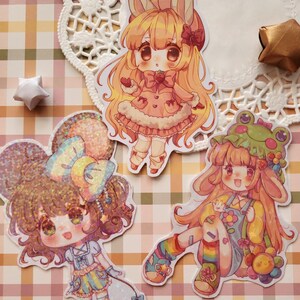 May include: Three colorful stickers featuring anime-style characters. One sticker depicts a girl with bunny ears in a pink dress. Another shows a girl with a large bow and stars. The third sticker shows a girl with a frog hat and rainbow socks.