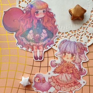 Puede incluir: Dos pegatinas con personajes de estilo anime con ojos grandes y cabello colorido. Una pegatina representa a una niña con un vestido verde, la otra a una niña con un vestido rosa con una oveja. También se ve una estrella dorada y una estrella blanca.