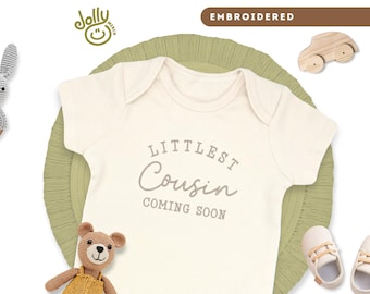 May include: Cream-colored baby onesie with the embroidered text "Littlest Cousin Coming Soon." The onesie is surrounded by baby toys, including a crochet bunny, a teddy bear, and a pair of baby shoes. The word "EMBROIDERED" is in the top right corner.