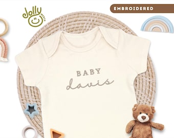 May include: Cream-colored baby onesie with the name "Davis" embroidered in a cursive font. The word "BABY" is embroidered above the name. The image includes a teddy bear, wooden toys, and the text "EMBROIDERED".