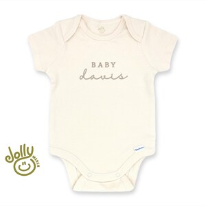 Pu&ograve; includere: Body da bambino color crema con maniche corte e scollo tondo. Il testo "BABY davis" &egrave; stampato in grigio sul petto. Un piccolo logo &egrave; visibile in basso. Il logo Jolly Merch si trova in basso a sinistra.