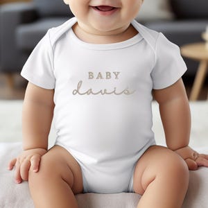 Pu&ograve; includere: Un body da bambino bianco con il nome "Davis" in corsivo e la parola "BABY" sopra. Il testo &egrave; di colore marrone chiaro. Il body &egrave; a maniche corte e ha uno scollo rotondo.