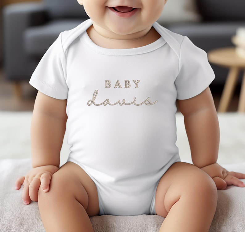 Pu&ograve; includere: Un body da bambino bianco con il nome "Davis" in corsivo e la parola "BABY" sopra. Il testo &egrave; di colore marrone chiaro. Il body &egrave; a maniche corte e ha uno scollo rotondo.