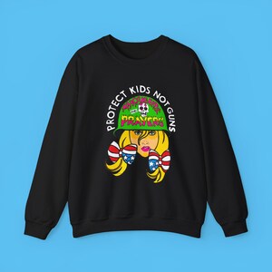 May include: Black sweatshirt with a graphic print of a woman wearing a green helmet with the text "Protect Kids Not Guns" and "Thoughts and Prayers" in pink and green letters.