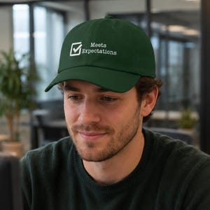 May include: A dark green baseball cap featuring a white embroidered graphic of a square checkbox with a checkmark next to the text "Meets Expectations" in a clean, sans-serif font centred on the front panel of the hat.