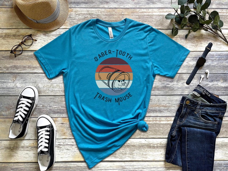 May include: A turquoise blue t-shirt with a retro-style graphic of a trash can with a mouse inside. The graphic says "Saber-Tooth Trash Mouse".
