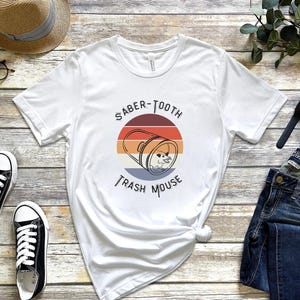 May include: White t-shirt with a retro-style graphic of a trash can with the text "Saber-Tooth Trash Mouse".