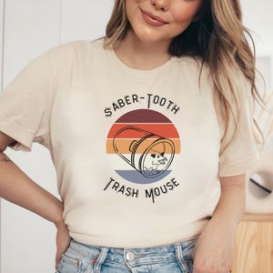 May include: A beige t-shirt with a retro-style graphic design featuring a trash can with a mouse inside and the text "Saber-Tooth Trash Mouse".