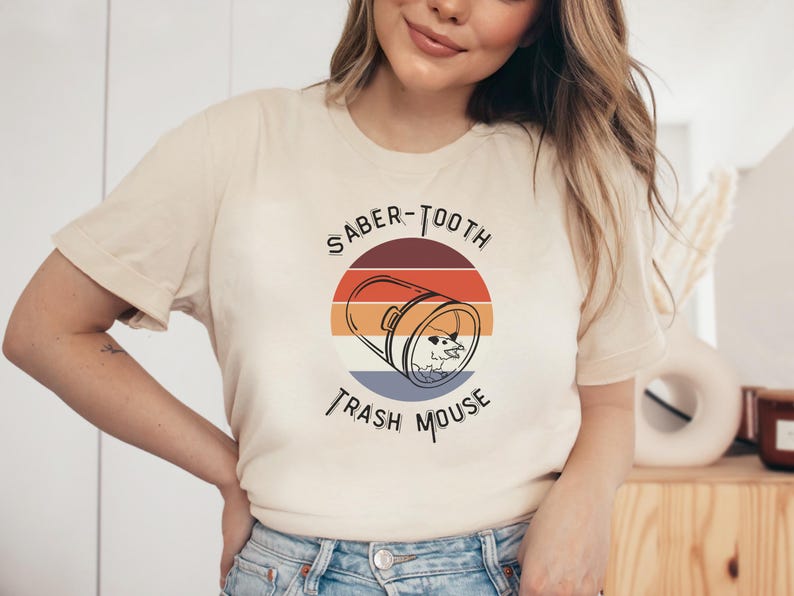 May include: A beige t-shirt with a retro-style graphic design featuring a trash can with a mouse inside and the text "Saber-Tooth Trash Mouse".