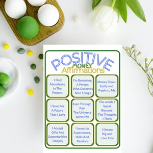 Puede incluir: Un fondo blanco con una bandeja de madera que contiene esferas blancas y verdes. Una tarjeta con el texto "POSITIVE MONEY Affirmations" y afirmaciones como "I Find Abundance In The Present" está en el centro.
