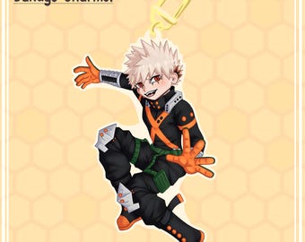 Op de afbeelding: Een cartoon-personage hanger met een zwart en oranje kostuum, blond stekelig haar en rode ogen. Het personage staat in een springende pose met één arm omhoog en de andere arm gestrekt. De hanger heeft een gouden metalen ring om aan een sleutelhanger of tas te bevestigen.