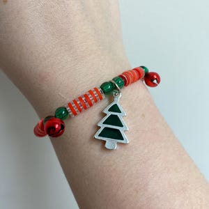 K&ouml;nnte beinhalten: Ein rotes und gr&uuml;nes Perlenarmband mit einem silbernen Anh&auml;nger in Form eines Weihnachtsbaums. Das Armband hat kleine Gl&ouml;ckchen.