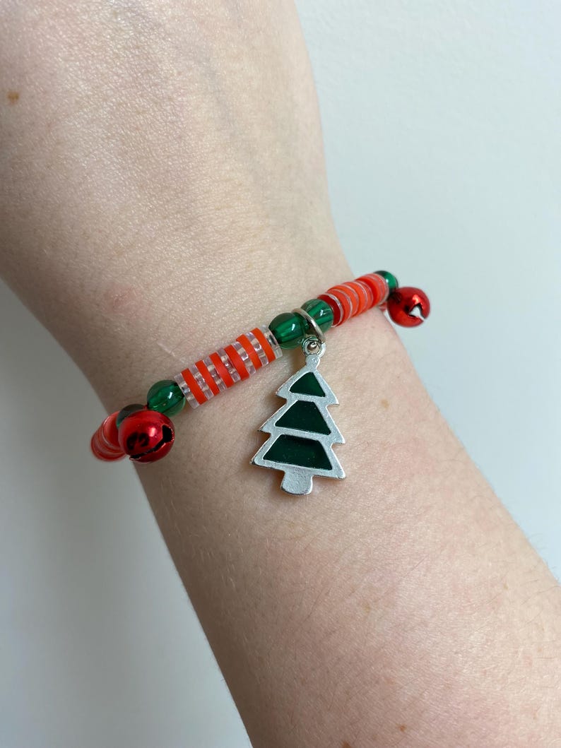 K&ouml;nnte beinhalten: Ein rotes und gr&uuml;nes Perlenarmband mit einem silbernen Anh&auml;nger in Form eines Weihnachtsbaums. Das Armband hat kleine Gl&ouml;ckchen.
