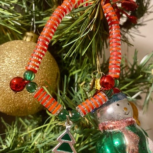 K&ouml;nnte beinhalten: Ein rot-wei&szlig; gestreifter Weihnachtsbaumschmuck aus Perlen mit einem gr&uuml;nen und silbernen Weihnachtsbaum-Anh&auml;nger. Der Schmuck h&auml;ngt an einem gr&uuml;nen Weihnachtsbaum.