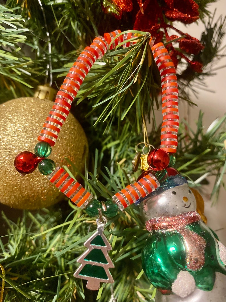 K&ouml;nnte beinhalten: Ein rot-wei&szlig; gestreifter Weihnachtsbaumschmuck aus Perlen mit einem gr&uuml;nen und silbernen Weihnachtsbaum-Anh&auml;nger. Der Schmuck h&auml;ngt an einem gr&uuml;nen Weihnachtsbaum.
