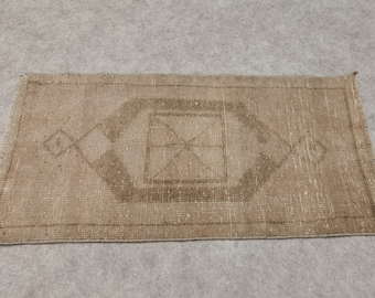 May include: A rectangular, beige rug with a geometric design. The rug features a central diamond-shaped pattern with an X inside, surrounded by a lighter beige field. The edges have a fringed detail.