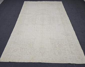 Könnte beinhalten: Ein rechteckiger, cremefarbener Teppich mit einem verblichenen, abgenutzten Aussehen. Der Teppich hat ein zentrales Medaillon-Design und sich wiederholende Muster in einem helleren Farbton. Der Teppich liegt auf einer dunkelgrauen Oberfläche.