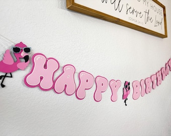 May include: A pink and white "Happy Birthday" banner with a flamingo wearing sunglasses. The banner is strung across a white wall, with a wooden framed sign in the background.