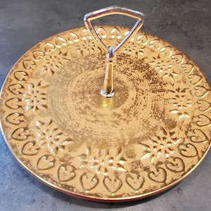 May include: A gold-colored ceramic serving tray with a decorative heart and flower pattern around the edge. The tray has a metal handle in the center.