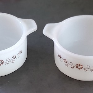 May include: Two white ceramic ramekins with brown floral designs. The ramekins have two handles on the sides.