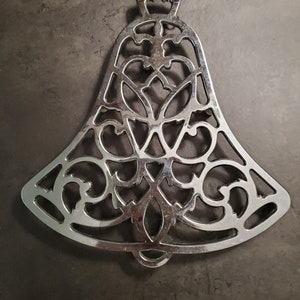 May include: A silver metal bell-shaped trivet with an intricate, ornate design. The trivet is a decorative and functional kitchen accessory.