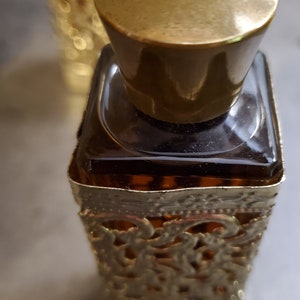 May include: A gold-colored bottle with a decorative, ornate design. The bottle is filled with a dark liquid and has a gold-colored cap.