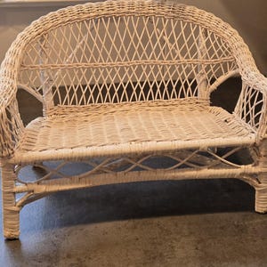 May include: A cream-colored wicker loveseat with a curved back and armrests. The woven design features a diamond pattern on the back and seat. The legs are sturdy and the overall design is classic and inviting.