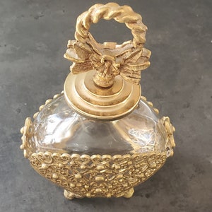 May include: A gold-toned glass bottle with a decorative, ornate lid and a gold-toned, intricate base. The lid features a twisted rope design and a bow.