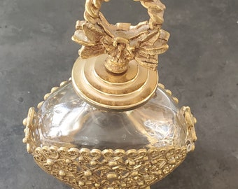 May include: A gold-toned glass bottle with a decorative, ornate lid and a gold-toned, intricate base. The lid features a twisted rope design and a bow.