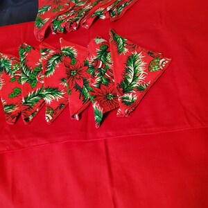 May include: A set of red Christmas napkins with a green poinsettia and holly leaf design. The napkins are folded and stacked on a red tablecloth.
