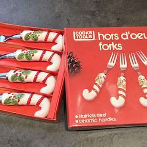 May include: Set of four hors d'oeuvre forks with stainless steel tines and ceramic handles. The handles are white with red stripes and a holly design. The red box reads "Cooks Tools" and "hors d'oeuvre forks".