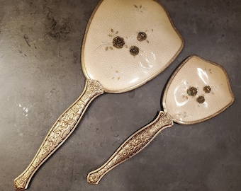 May include: A set of two vintage hand mirrors with gold handles and floral designs. The larger mirror has a white background with gold floral accents and the smaller mirror has a white background with gold floral accents and a gold trim.