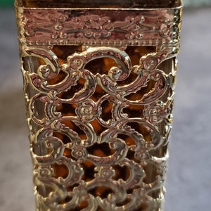 May include: A gold and brown glass bottle with a decorative gold filigree design. The bottle has a square shape and a gold cap.