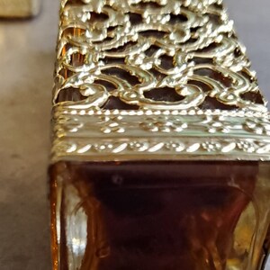 May include: A gold-colored glass bottle with a decorative, intricate design. The bottle is filled with a dark liquid.