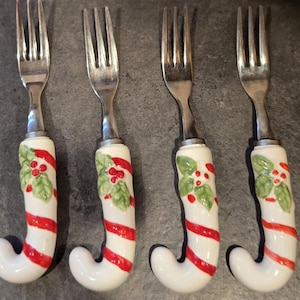 May include: Four silver forks with candy cane-shaped handles. The handles are white with red stripes and holly berry accents. The forks are arranged in a row, perfect for holiday entertaining.