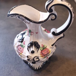 May include: A white ceramic pitcher with a black handle and floral design. The pitcher has a black rim and a black floral pattern with pink, yellow, and green accents.