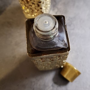 May include: A gold-colored glass bottle with a clear glass stopper. The bottle has a textured surface and is filled with a dark liquid.
