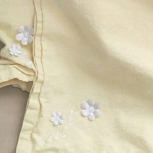 May include: A pale yellow fabric with white embroidered flowers. The flowers are simple and delicate, with a few petals each.