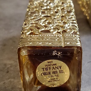 May include: A gold-colored glass bottle of Tiffany perfume with a gold label that reads "BQT PERFUME TIFFANY L'ARGENE PROD. DIST 2 FL. OZ COMP NEW YORK NY".
